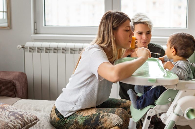 Family life. Lesbian couple with baby at home. One of them spoon feeding baby who is seated in a high chair in the living room