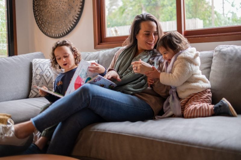 A mother laughs and plays with her two young children. They are snuggled up on the couch together and enjoying quality family time.